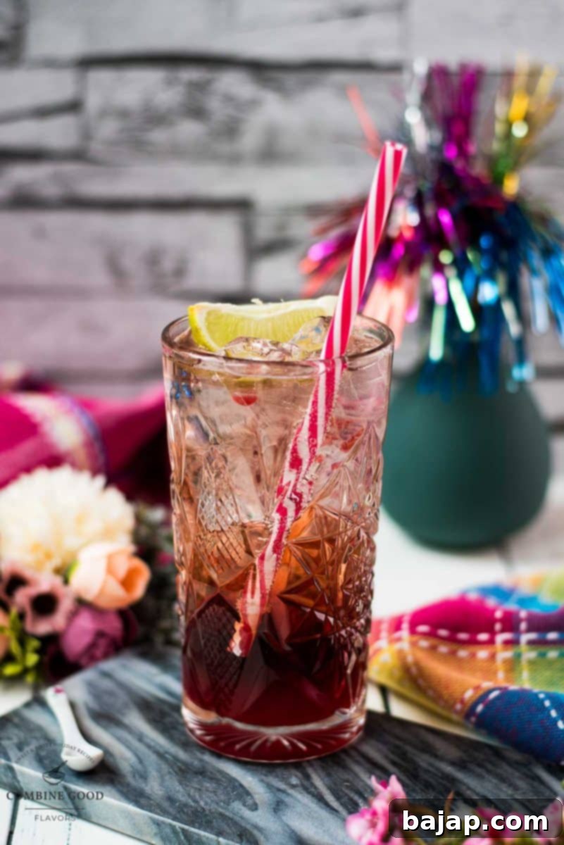 Gorgeous highball glass filled with raspberry gin tonic, using sloe gin. Garnished with a lime wedge and a drinking straw, against a light background.