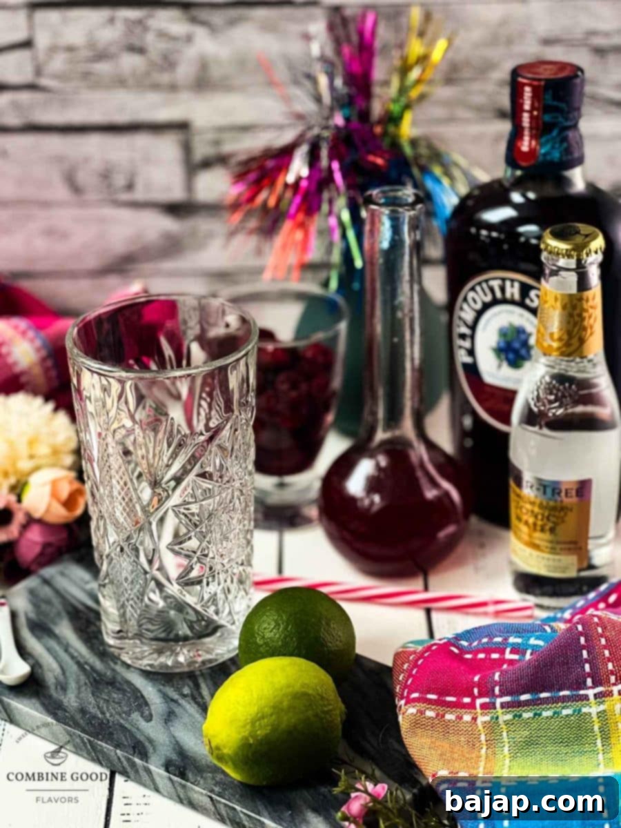 Ingredients for Raspberry Gin and Tonic: cubed ice, lime slices, tonic water, raspberry syrup, and a bottle of Plymouth sloe gin, arranged on a rustic surface.