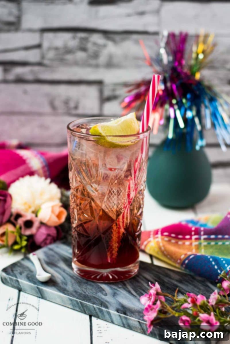Gorgeous highball glass filled with raspberry gin tonic, using sloe gin. Garnished with a lime wedge and a drinking straw, against a slightly darker background.