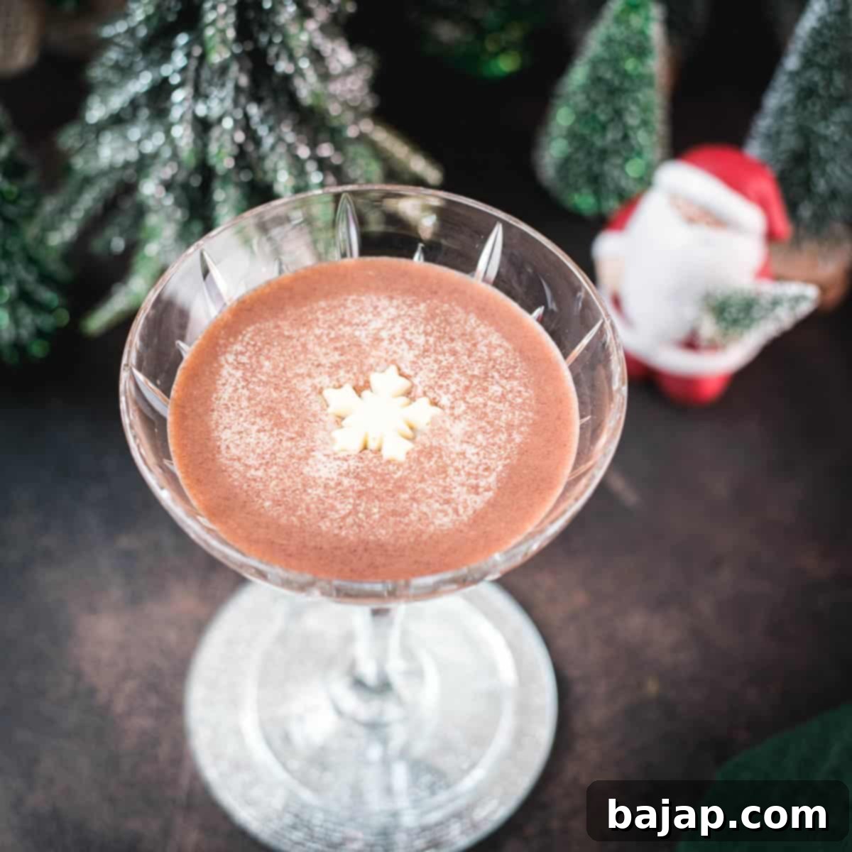 Festive chocolate martini with a white chocolate snowflake garnish, placed on mirrored coaster next to a Santa clause figurine.