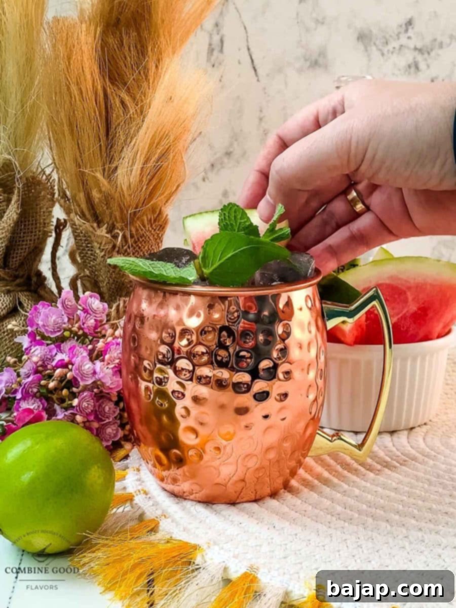 A hand carefully placing a fresh watermelon slice and mint sprig into the copper mug as a beautiful and aromatic garnish for the Watermelon Moscow Mule.
