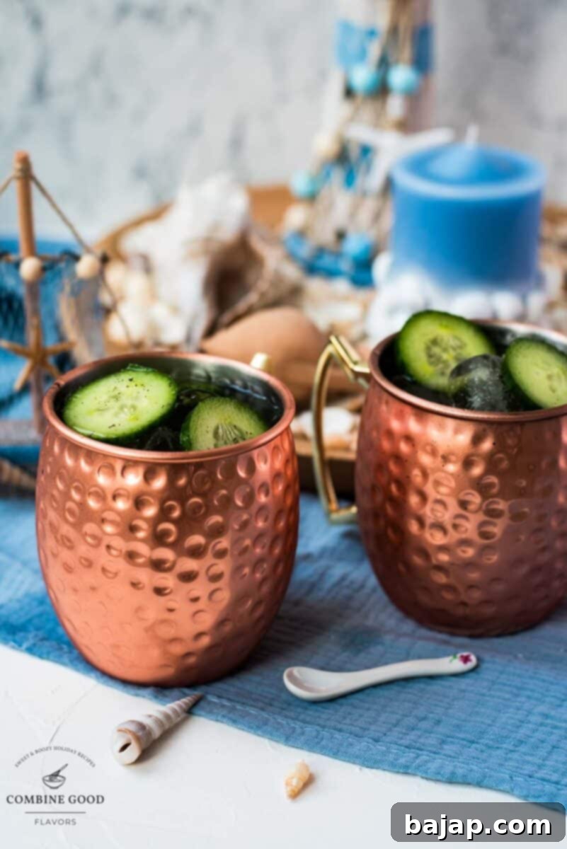 A refreshing mint cucumber Moscow mule in a frosted copper mug, garnished with cucumber slices and a lime wedge, sitting on a white and blue surface.