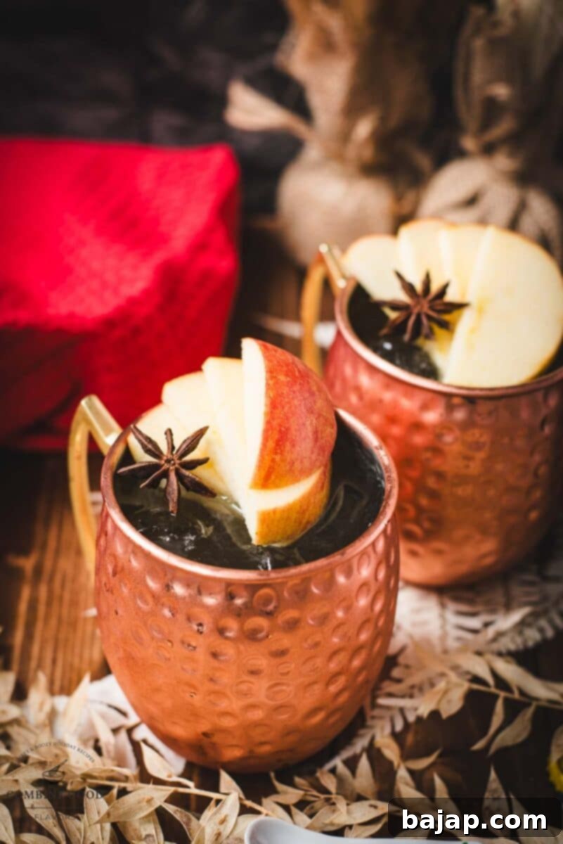 Autumn Apple Cider Moscow Mule 2 Copper mug, filled with delicious apple cider Moscow mule, garnished with fresh apple slices and a star anise. Placed on wooden board, next to a red cloth.