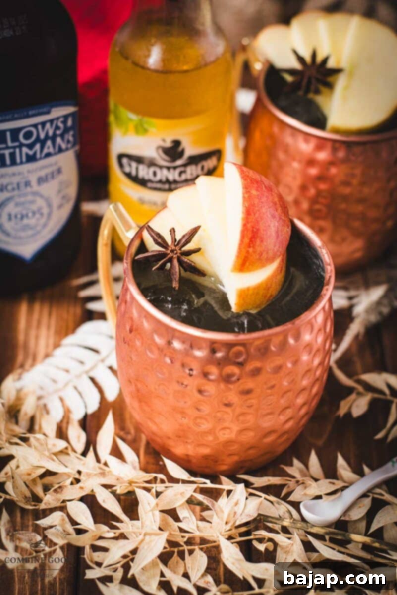 Crisp Apple Mule 11 Copper mug, filled with delicious apple cider Moscow mule, garnished with fresh apple slices and a star anise. Placed on wooden board, next to a red cloth.