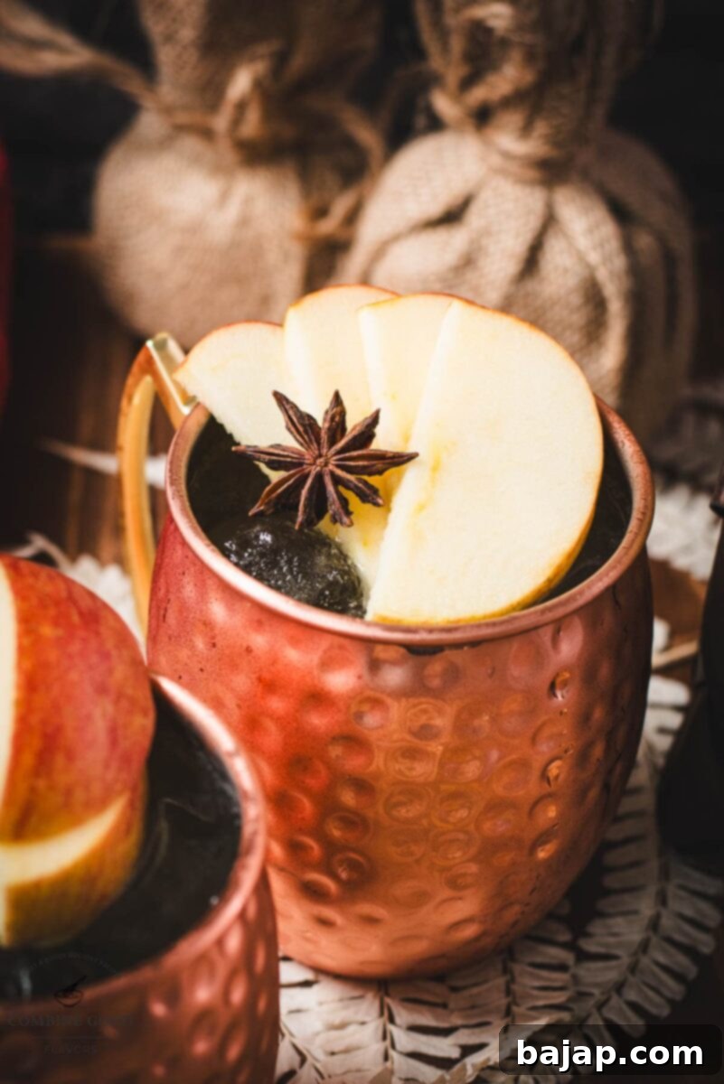 Crisp Apple Mule 12 Copper mug, filled with delicious apple cider Moscow mule, garnished with fresh apple slices and a star anise. Placed on wooden board.