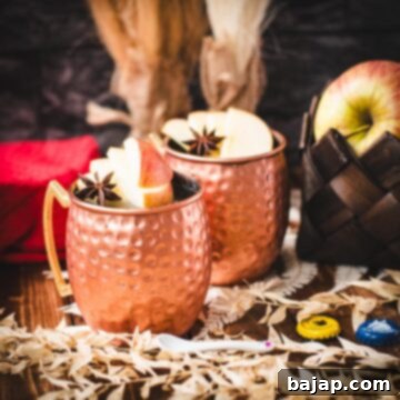 Copper mug, filled with delicious apple cider Moscow mule, garnished with fresh apple slices and a star anise. Placed on wooden board, next to a red cloth.