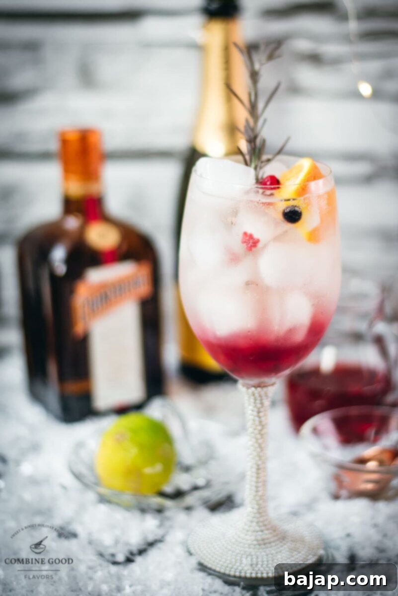 A stunning layered Prosecco Cosmo cocktail in a wine glass, garnished with a sprig of fresh rosemary, a bright orange slice, and ruby red and black currants.