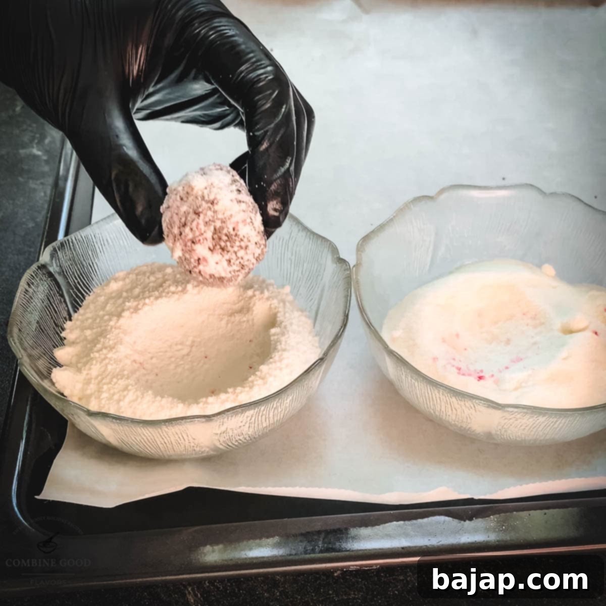 Hand with black gloves holding dough ball over a bowl filled with powdered sugar.