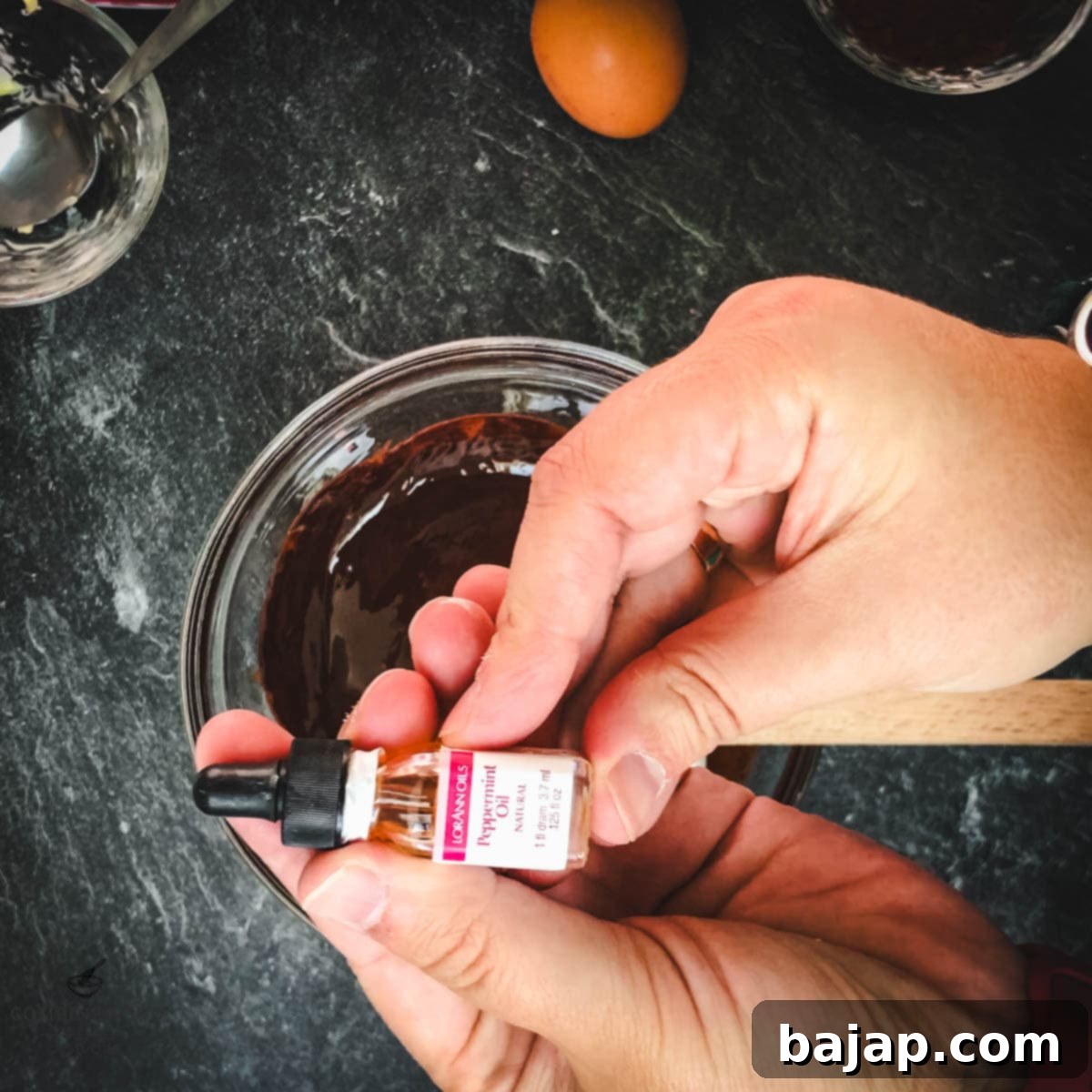 Hands holding peppermint oil over a bowl of melted chocolate.