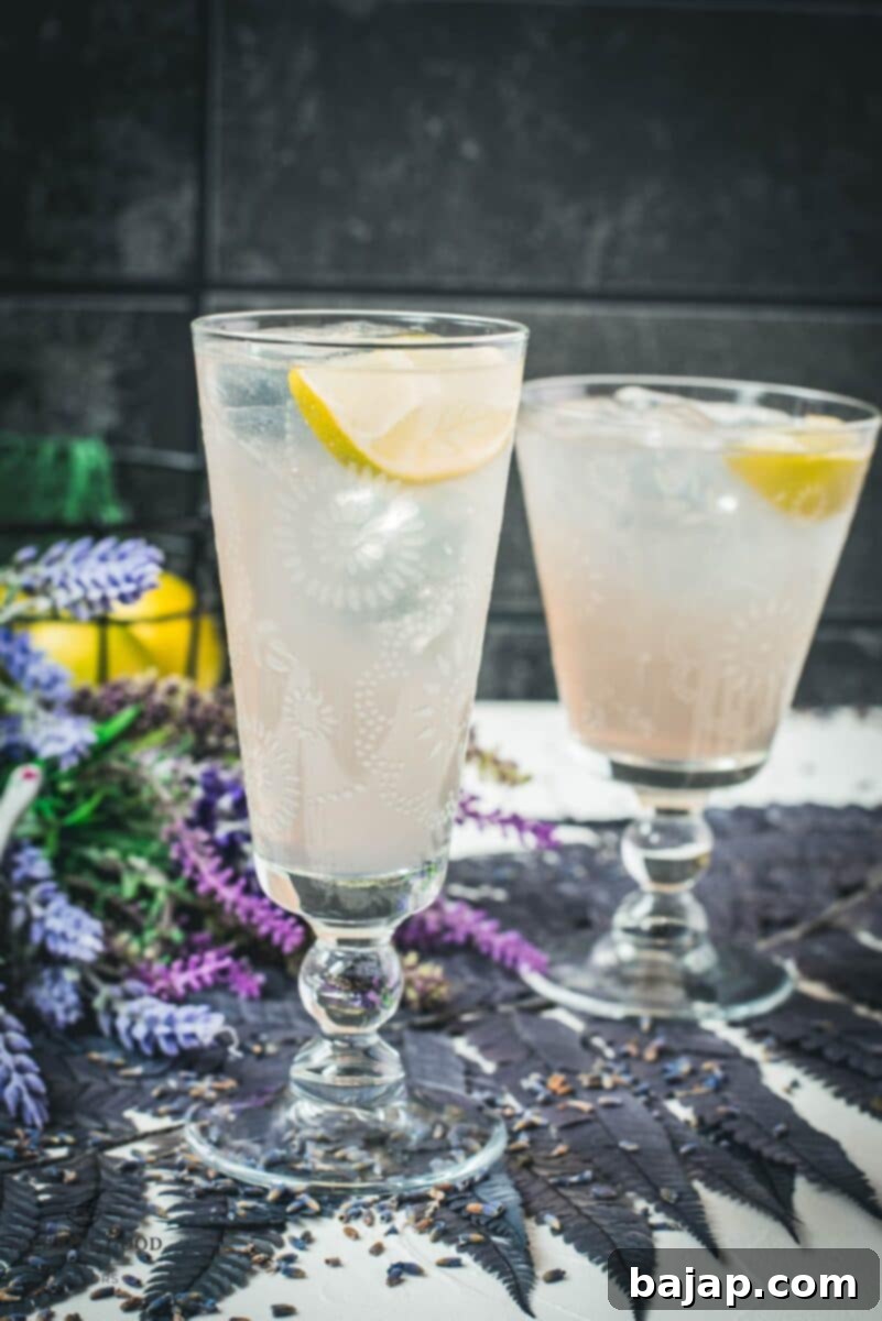 Two beautiful glasses with flower engraving filled with lavender Moscow Mule, arranged on dark blue purple leaves.