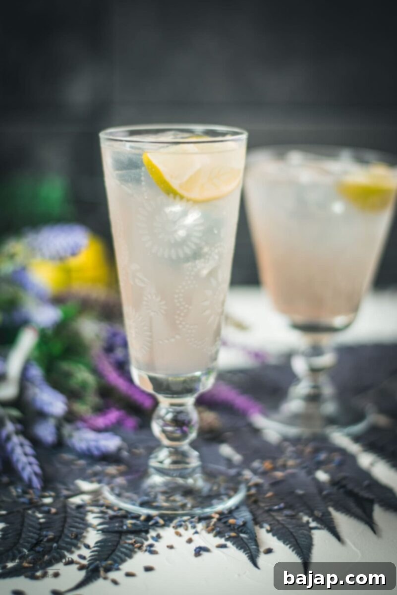 Two beautiful glasses with flower engraving filled with lavender Moscow Mule, arranged on dark blue purple leaves.