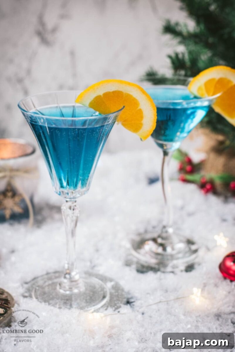 Gorgeous cocktail glasses filled with a stunning blue Christmas cocktail, garnished with an orange slice. Placed on mirrored coasters on snowy ground.