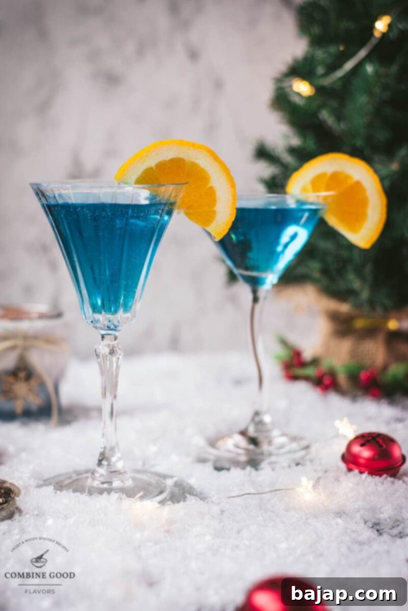 Gorgeous cocktail glasses filled with a stunning blue Christmas cocktail, garnished with an orange slice. Placed on mirrored coasters on snowy ground.
