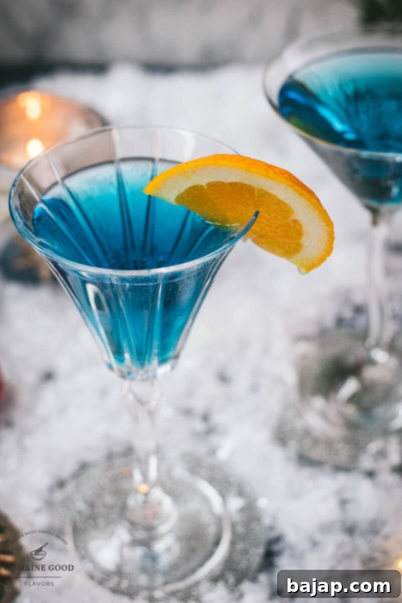 Gorgeous cocktail glasses filled with a stunning blue Christmas cocktail, garnished with an orange slice. Placed on mirrored coasters on snowy ground.