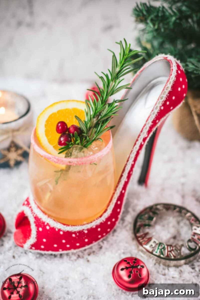 Festive vodka cranberry spritzer, garnished with rosemary twig, orange slice, and cranberries, served in a glass that is placed in a christmassy red high heel, with additional garnishes arranged artfully beside it.