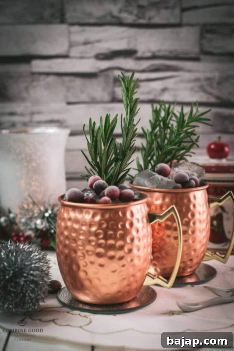 Two copper mugs filled with a delicious cranberry Moscow mule, placed on mirrored coaster and garnished with rosemary twigs and frozen cranberries.
