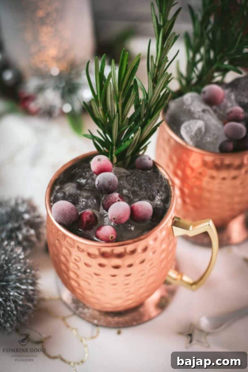 Two copper mugs filled with a delicious cranberry Moscow mule, placed on mirrored coaster and garnished with rosemary twigs and frozen cranberries.