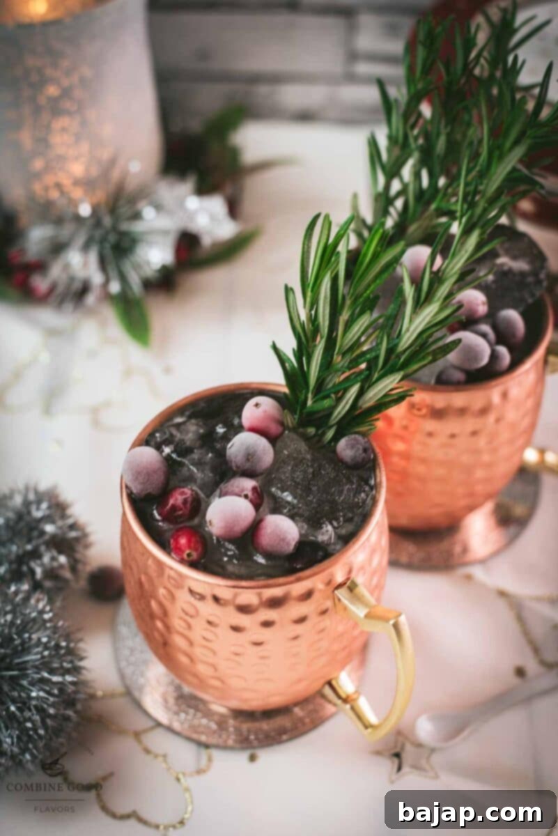 Two copper mugs filled with a delicious cranberry Moscow mule, placed on mirrored coaster and garnished with rosemary twigs and frozen cranberries.