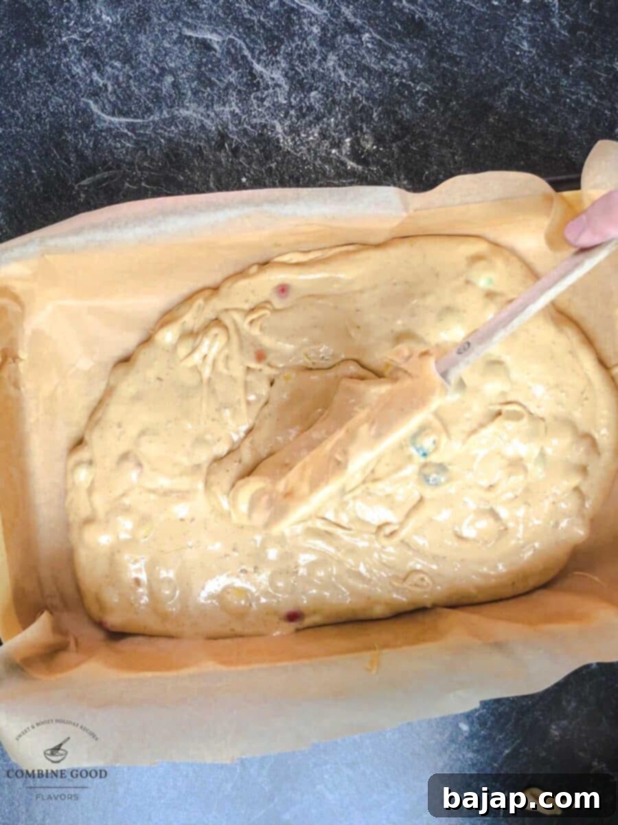 M&M cookie bar batter being spread evenly with a spatula into a parchment-lined baking pan.