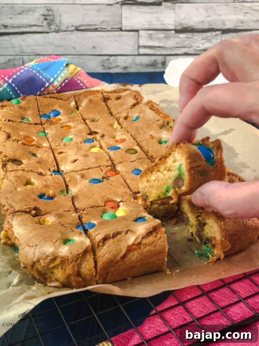 Colorful M and M Cookie Bars 7 A close-up of a hand gently picking up a freshly sliced M&M cookie bar from a tray.