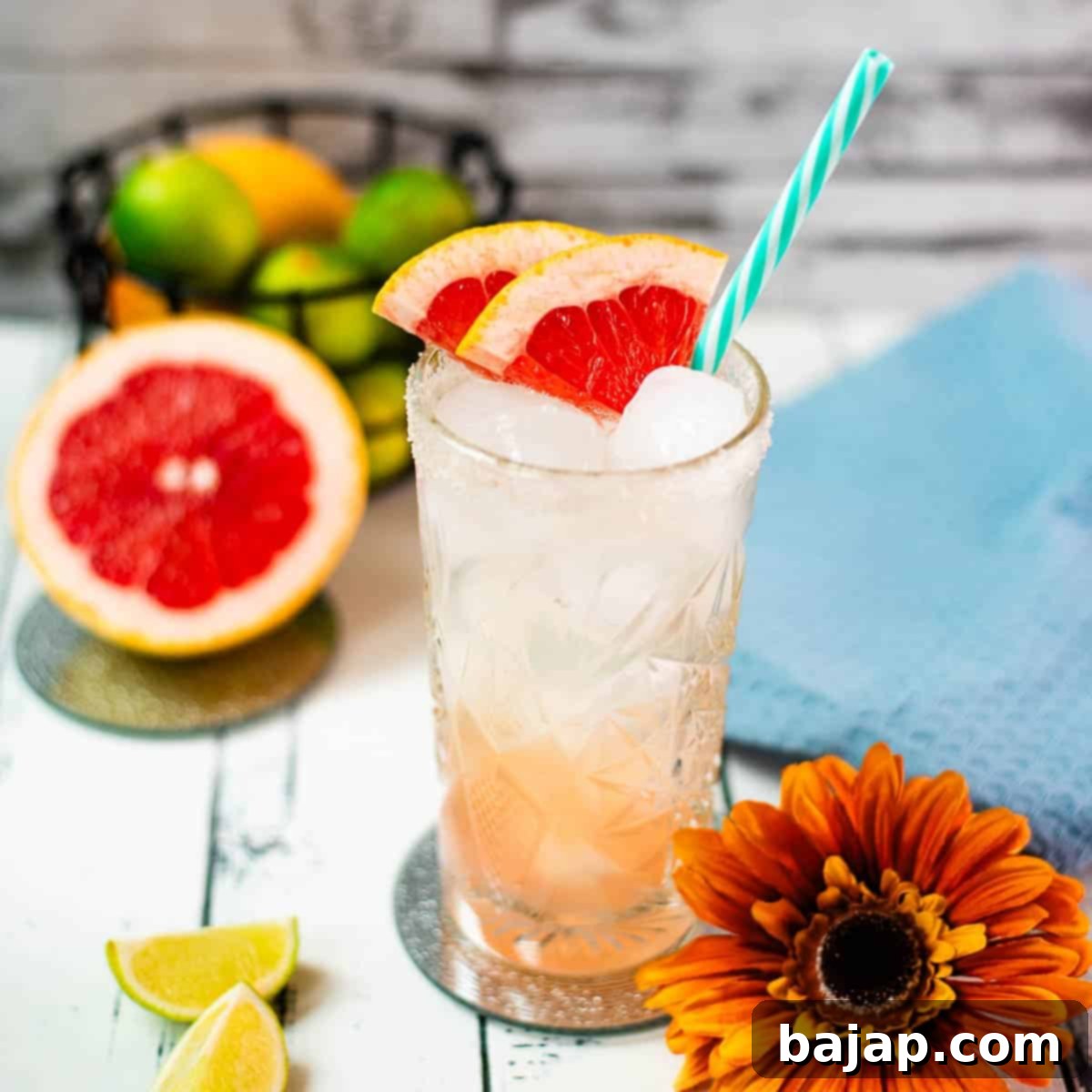 Colorful and refreshing paloma cocktail ina a highball glass, placed on mirrored coaster. Garnished with grapefruit slices and a straw.