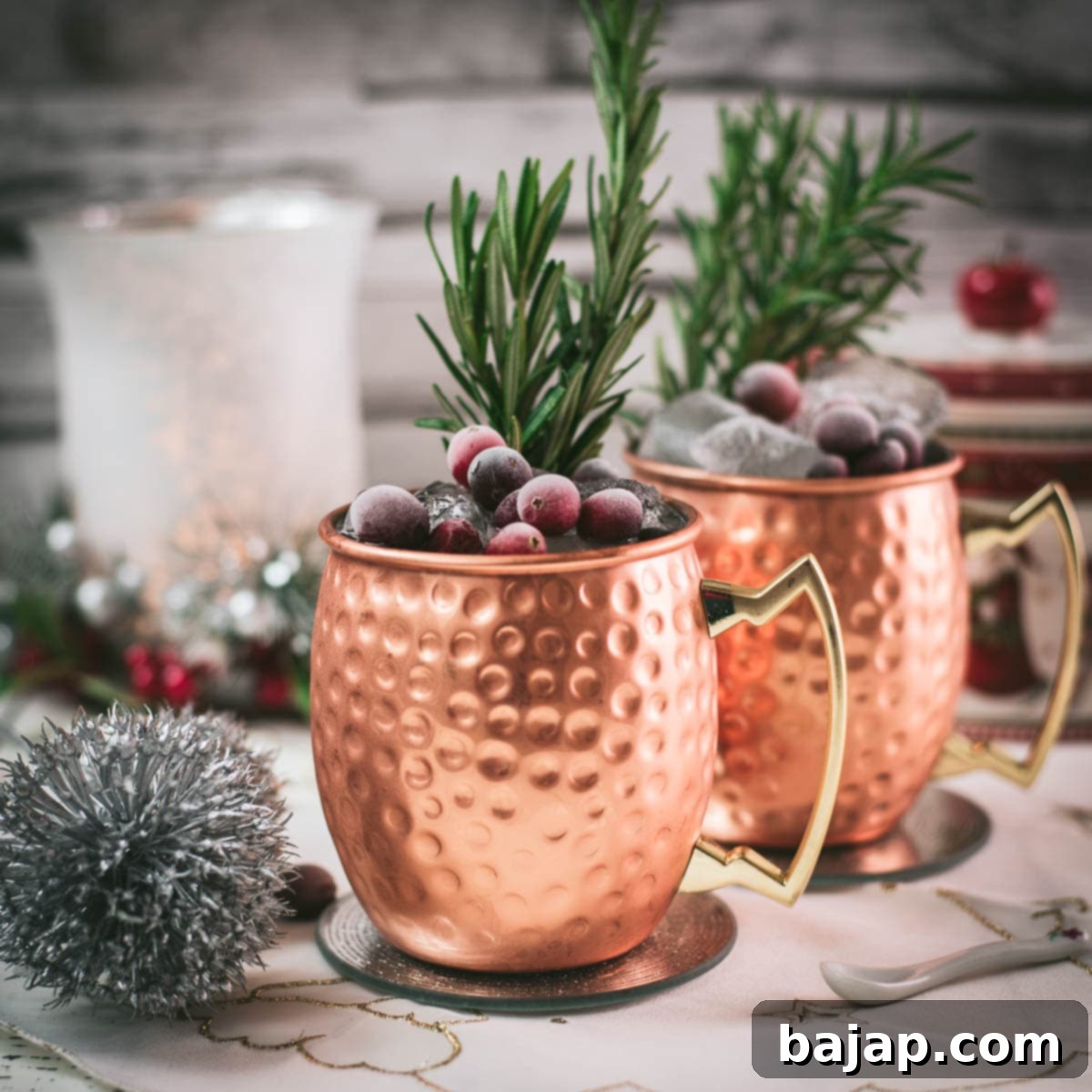 Two copper mugs filled with a delicious cranberry Moscow mule, placed on mirrored coaster and garnished with rosemary twigs and frozen cranberries.
