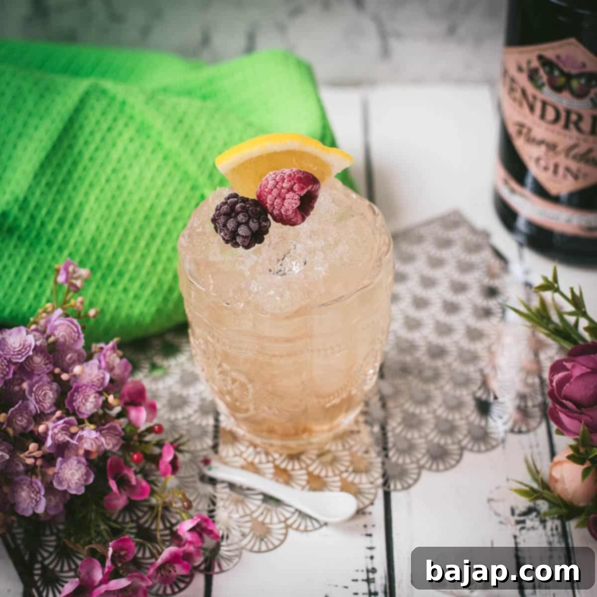 Fancy bramble cocktail in gorgeous glass, placed next to flowers and a white spoon. Garnished with a blackberry, raspberry, and a lemon wedge.