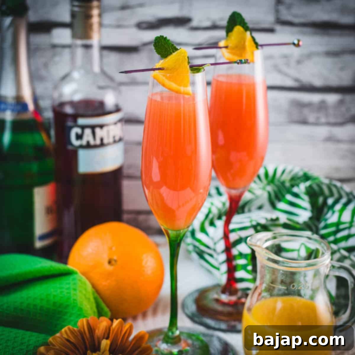 Two champagne glasses filled with vibrant Campari Mimosa. Garnished with an Orange slice and mint leaf on a cocktail skewer.