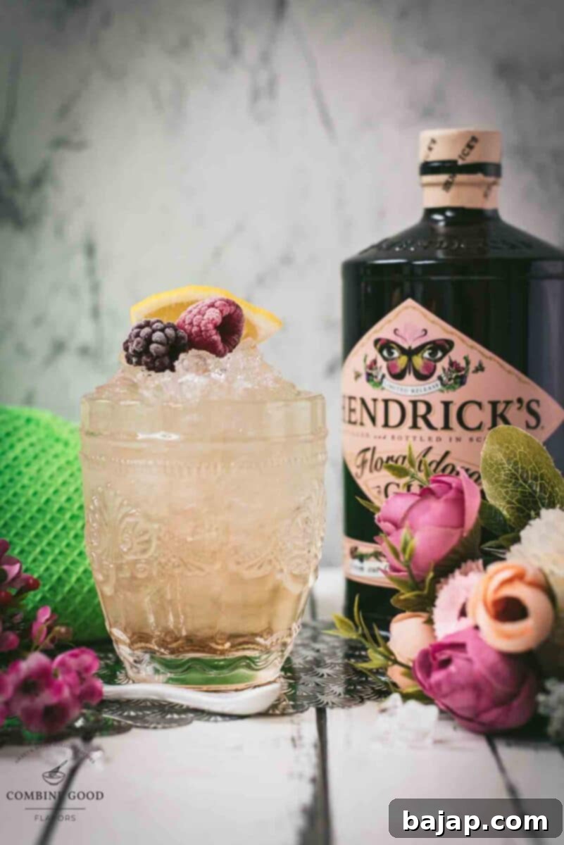 Fancy bramble cocktail in gorgeous glass, placed next to flowers and a white spoon. Garnished with a blackberry, raspberry, and a lemon wedge.