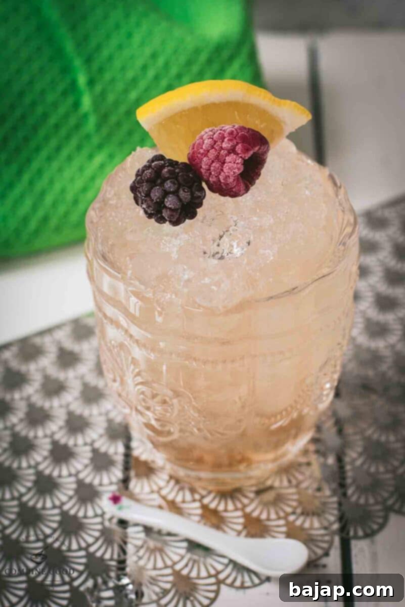 Fancy bramble cocktail in gorgeous glass, placed next to flowers and a white spoon. Garnished with a blackberry, raspberry, and a lemon wedge.