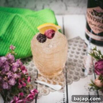 Fancy bramble cocktail in gorgeous glass, placed next to flowers and a white spoon. Garnished with a blackberry, raspberry, and a lemon wedge.