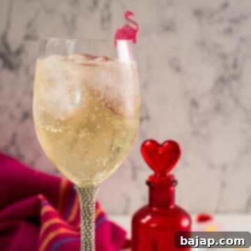 A beautiful Valentine's gin cocktail, garnished with a gorgeous flamingo cocktail pick, served in an elegant wine glass. It's set on a mirrored coaster next to a heart-shaped flask.
