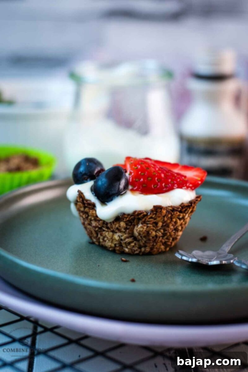 Crunchy Granola Bites 12 A close-up of a delightful little granola cup, expertly filled with creamy natural yogurt and generously topped with fresh, bright red strawberries, sweet blue grapes, and finished with a glistening drizzle of maple syrup. The cup is beautifully arranged on a dark green plate, highlighting its appetizing appearance.