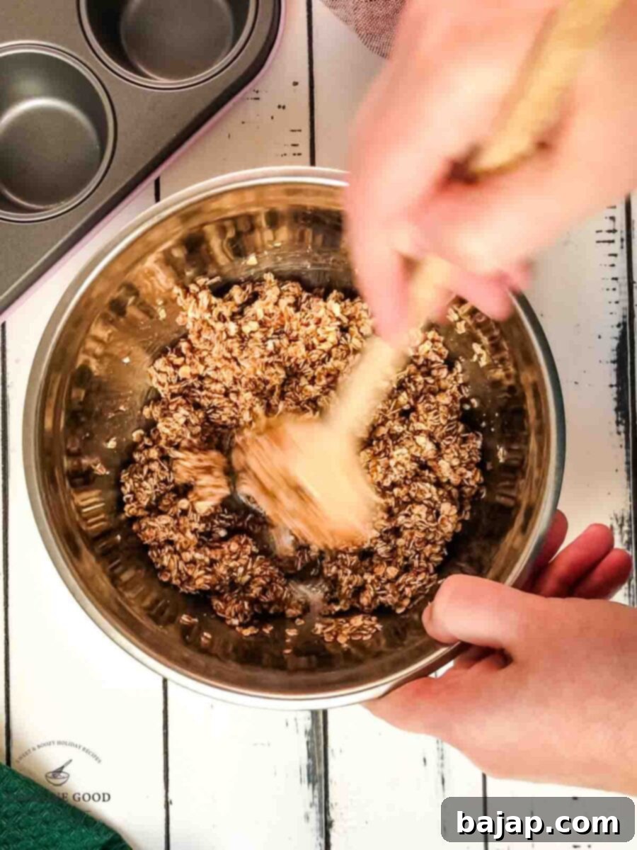 Crunchy Granola Bites 4 A hand is holding a clear glass mixing bowl, vigorously stirring a granola mixture with a wooden spoon. The mixture contains oats, cinnamon, vanilla, almond butter, salt, and agave syrup, ensuring everything is well coated.