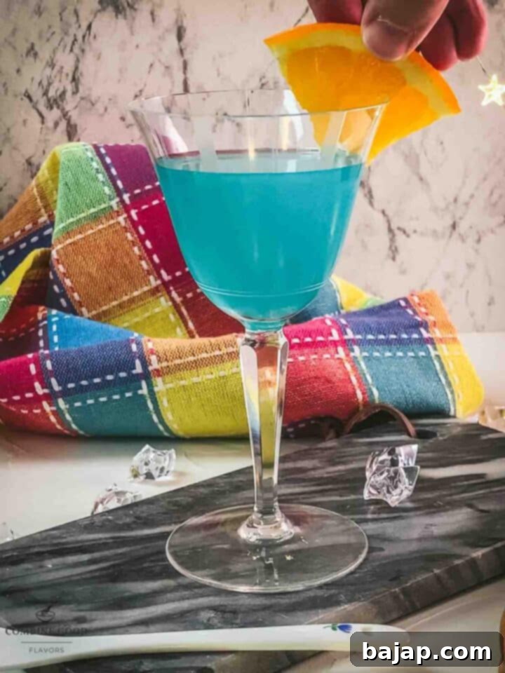 Hand placed orange slice on the brim of a cocktail glass filled with a blue lady cocktail.