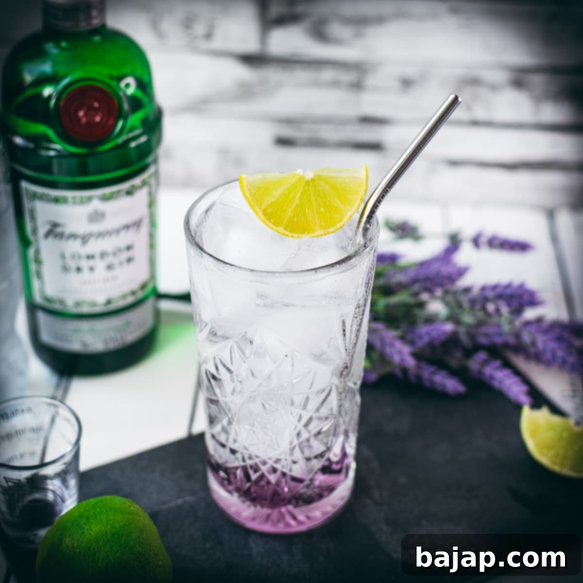 Highball glass filled with lavender gin and tonic, garnished with lime wedge and straw. Placed on black slate plate.