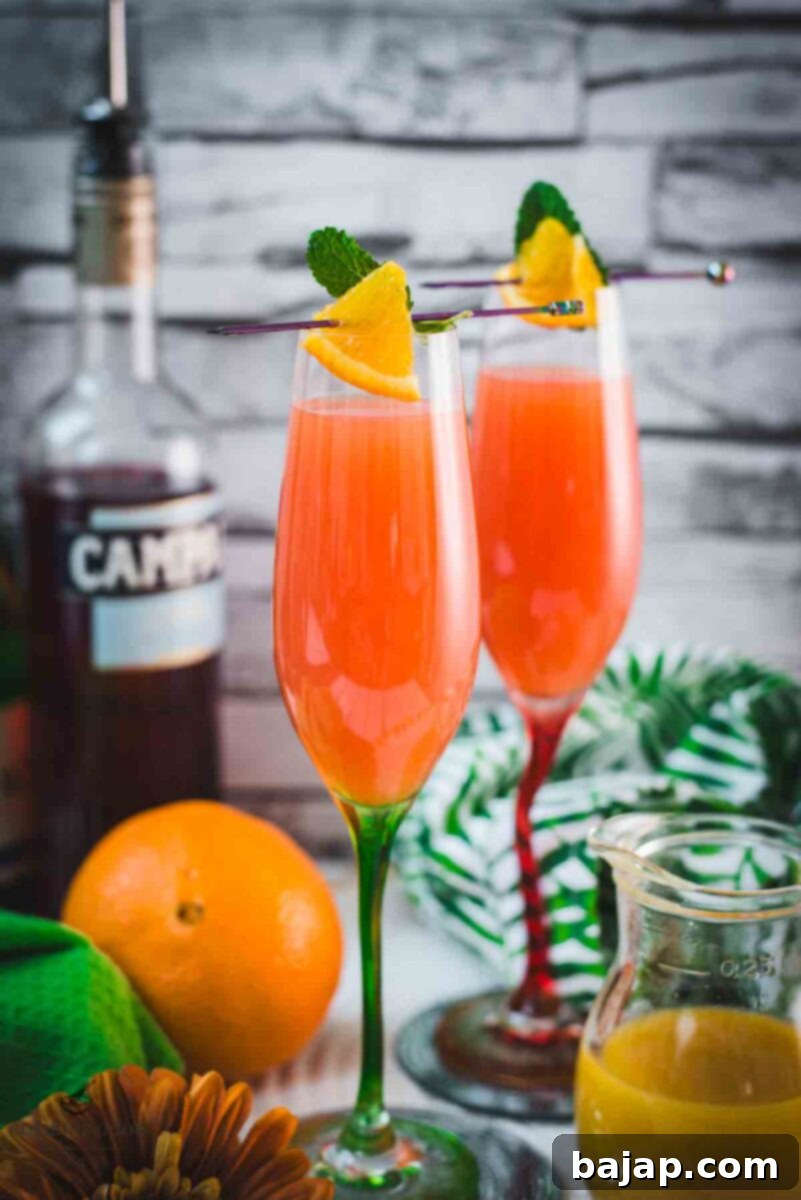 Two champagne glasses filled with vibrant Campari Mimosa, garnished with an orange slice and mint leaf on a cocktail skewer.