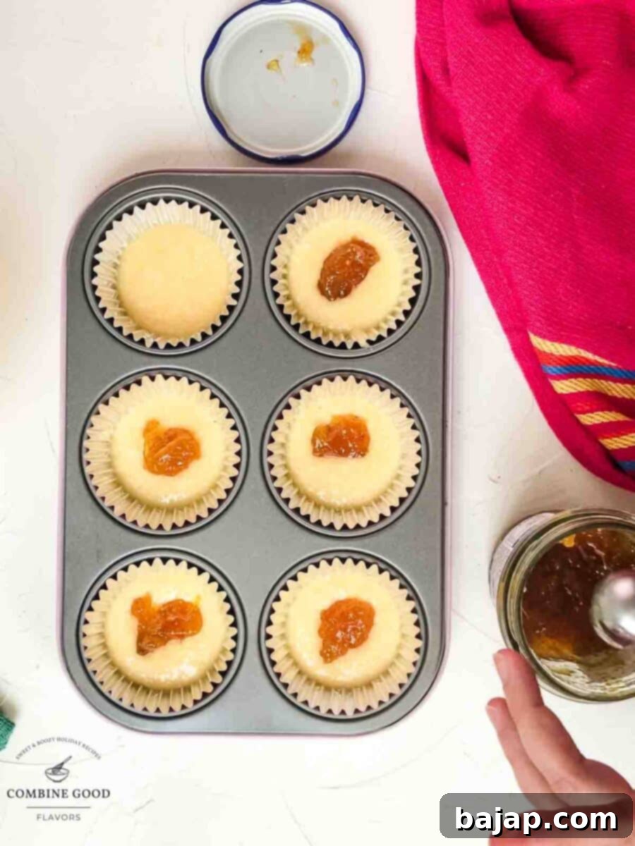 A teaspoon of fig jam being placed precisely into the center of each half-filled muffin cup.