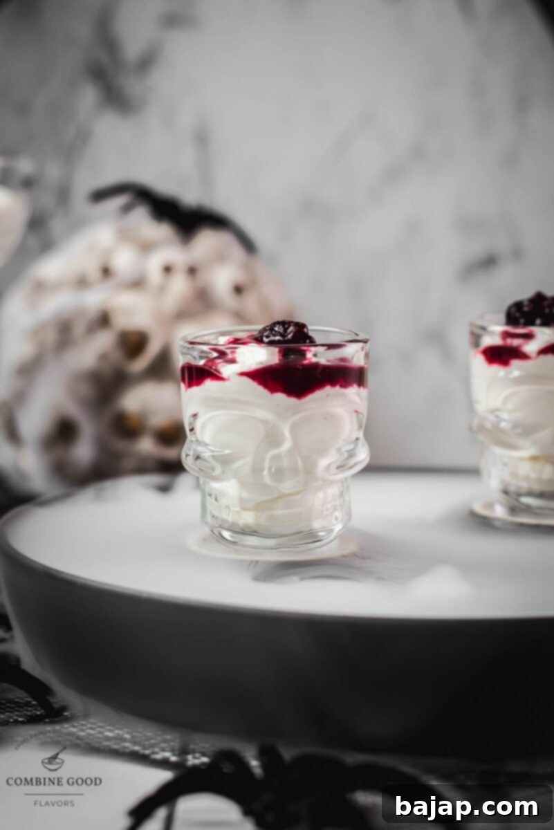 Spooky blood clot berry brains mousse, served in skull shaped shot glasses. The dessert features a white base with red berry swirls, resembling a brain, and is topped with extra red syrup for a gory effect. The skull glasses are placed on a dark surface, enhancing the eerie atmosphere.