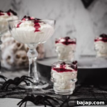 Spooky blood clot berry brains mousse, served in skull shaped shot glasses. A close-up of the mousse in a skull glass, showing the creamy texture, berry swirls, and a fresh drizzle of 'blood syrup'. The focus is on the dessert itself against a dark, blurred background.