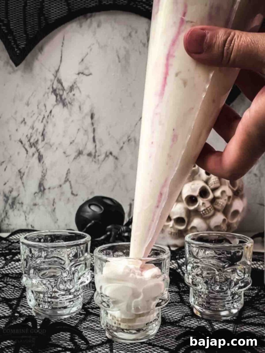 Pipe the mousse into the skull shaped shot glasses. A hand is holding a piping bag, carefully filling a small skull-shaped glass with white mousse. Other skull glasses are visible in the background, some already filled.