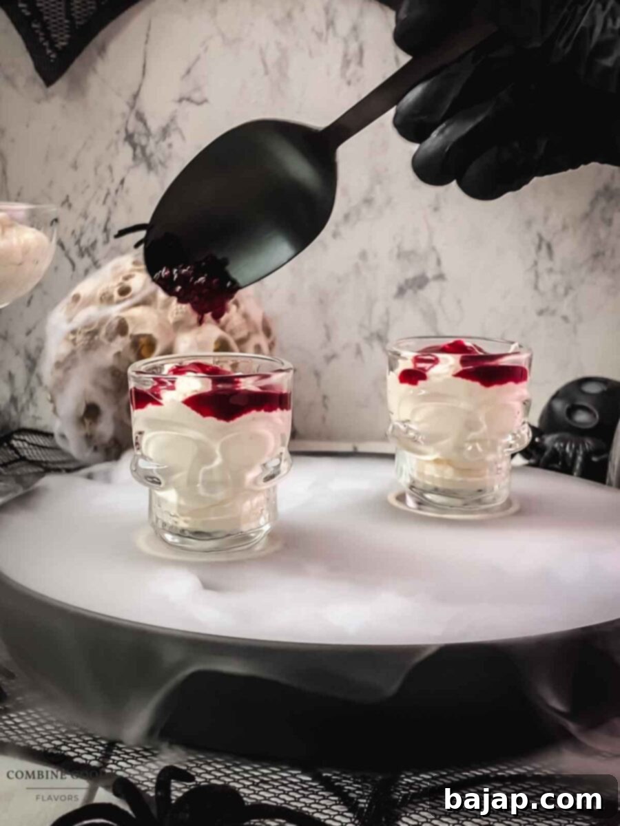 Top with the leftover strained pulp from the blood syrup production. A mousse in a skull-shaped glass is being topped with a spoonful of dark, fibrous berry pulp, enhancing the 'brain-like' and 'blood clot' texture. This highlights the sustainable aspect of using leftover fruit.