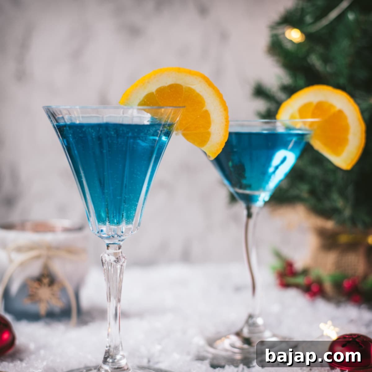 Gorgeous cocktail glasses filled with a stunning blue Christmas cocktail, garnished with an orange slice. Placed on mirrored coasters on snowy ground.