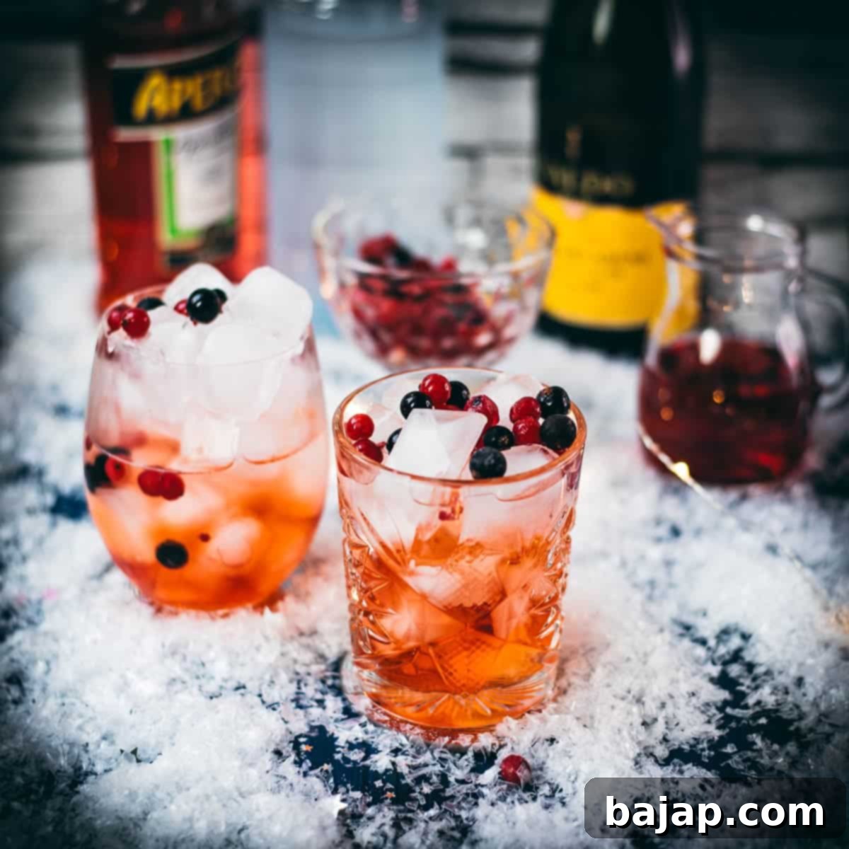 Two glasses filled with delicious cranberry Aaperol spritz, placed on snowy ground, garnished with a delicious berry mix.