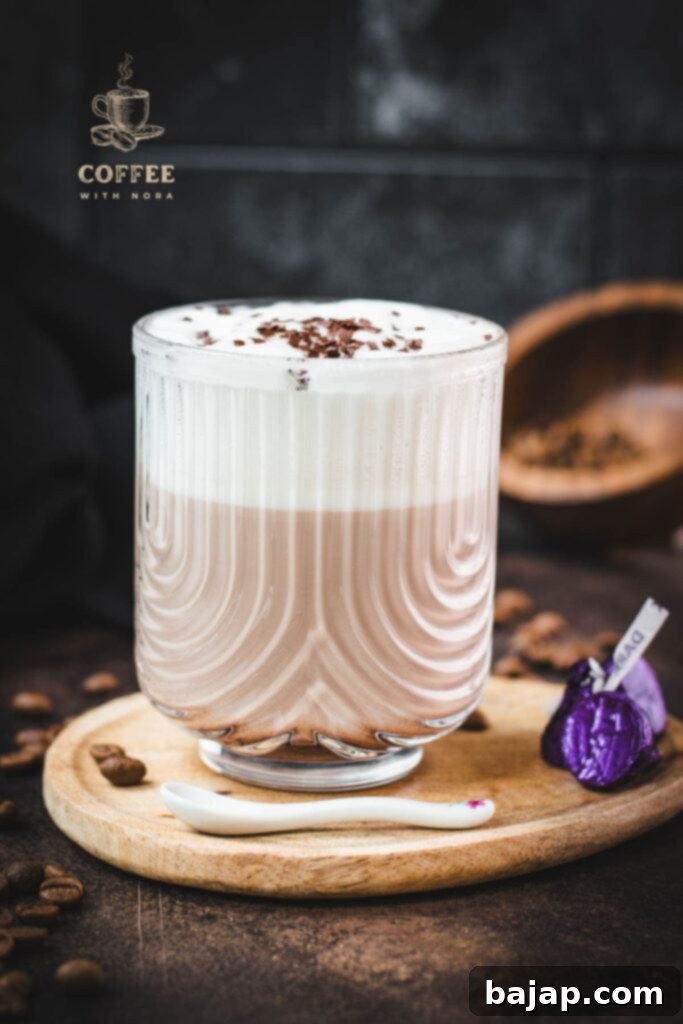 A beautifully crafted mocha in a glass, adorned with frothed milk and chocolate sprinkles, placed on a wooden plate with dark chocolate pieces.